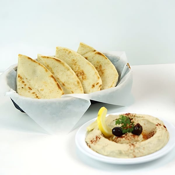 A basket of pita bread and a plate of hummus garnished with olives, lemon, and parsley.