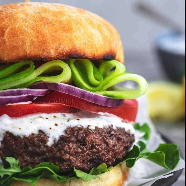 Close-up of a Juicy Gyros burger with a thick patty, tzatziki, tomato, red onion, and cucumber.