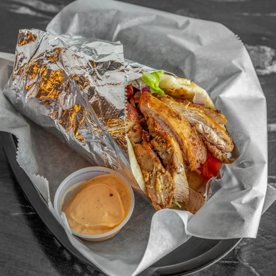 A Juicy Gyros chicken gyro, partially unwrapped from foil and paper, with a side of sauce.