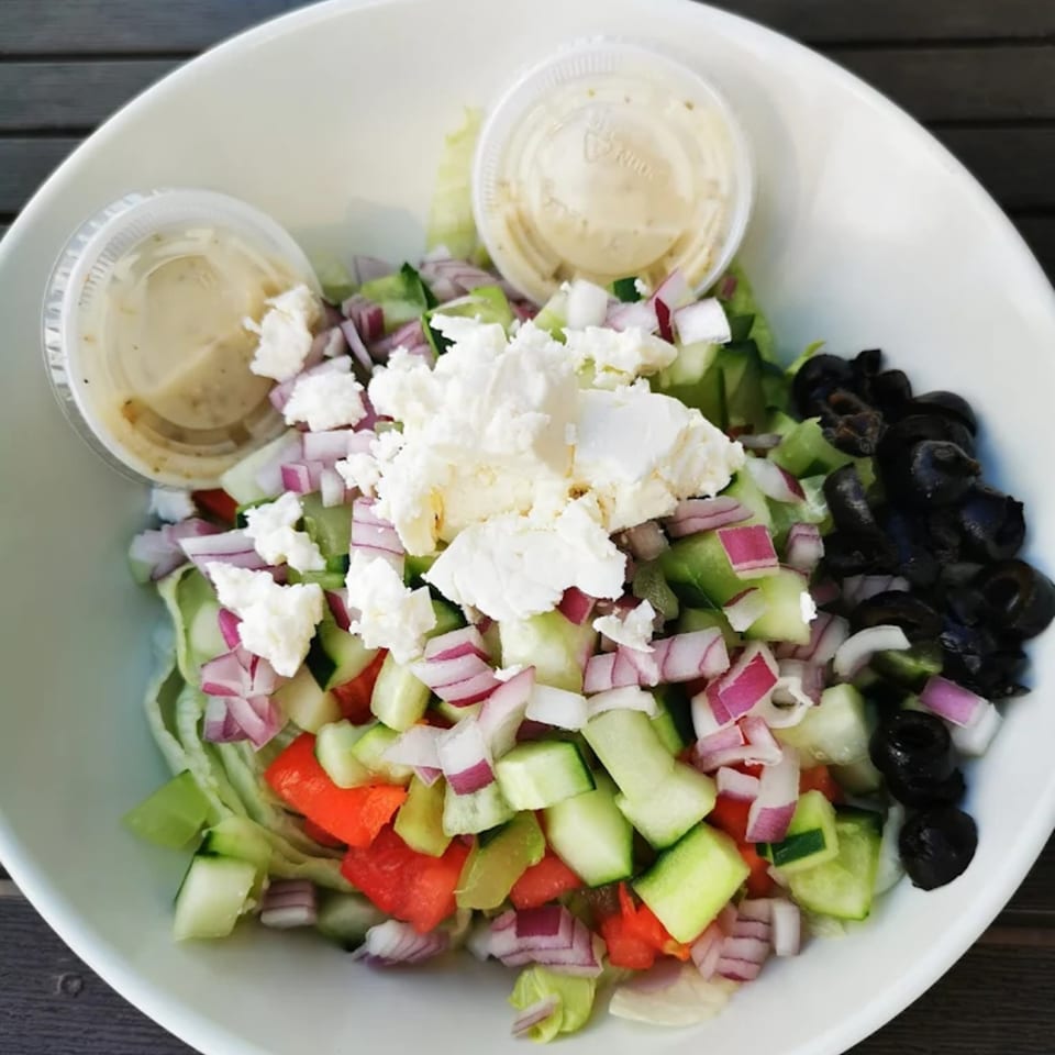 A Greek salad with feta, olives, red onion, cucumber, and bell pepper, with two dressing cups.